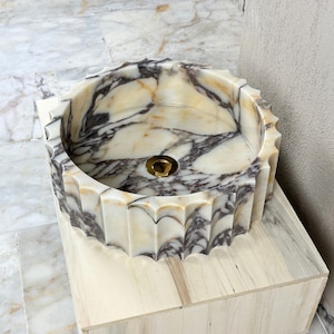 May include: A round marble sink with a scalloped edge. The sink features a cream and gray marbled pattern and a gold-colored drain. It is set on a light-colored wooden base. The sink's diameter is approximately 18 inches.