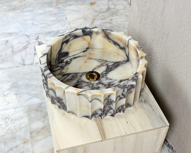 May include: A round marble sink with a scalloped edge. The sink features a cream and gray marbled pattern and a gold-colored drain. It is set on a light-colored wooden base. The sink's diameter is approximately 18 inches.
