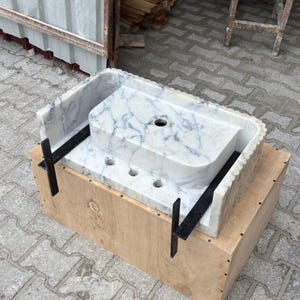 May include: A rectangular marble sink with gray veining, featuring a raised section with three faucet holes and a larger drain. The sink is set on a wooden crate and secured with black metal brackets. The sink's edges have a scalloped design.
