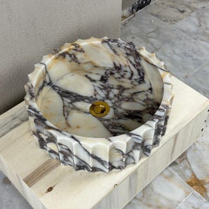 May include: A round marble sink with a scalloped edge. The sink features a gold-colored drain and a pattern of white, beige, and dark gray veining. The sink is set on a light-colored wooden base.