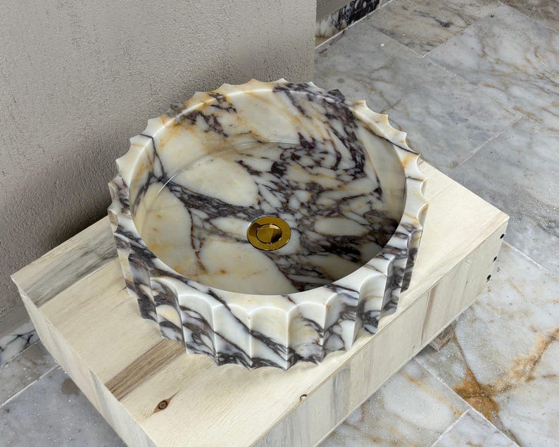 May include: A round marble sink with a scalloped edge. The sink features a gold-colored drain and a pattern of white, beige, and dark gray veining. The sink is set on a light-colored wooden base.