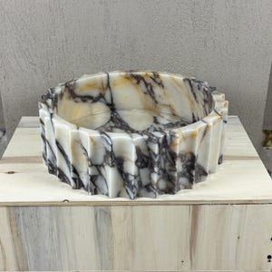 May include: A decorative, round marble basin with a fluted exterior. The basin features a cream and brown marbled pattern. The basin is sitting on a wooden box.