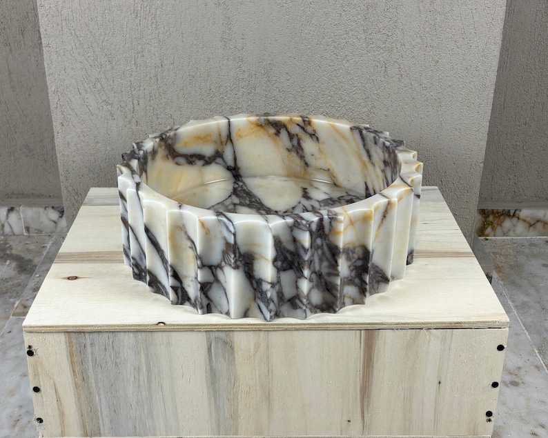 May include: A decorative, round marble basin with a fluted exterior. The basin features a cream and brown marbled pattern. The basin is sitting on a wooden box.