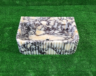 May include: Rectangular marble sink with a unique pattern of white, brown, and beige. The sink has a fluted exterior design and a drain hole. The sink is set against a green background.