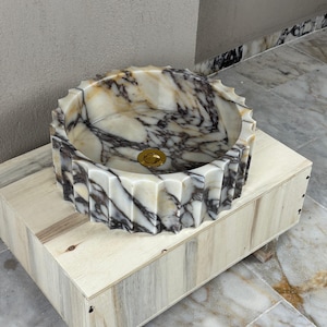 May include: A round marble sink with a decorative, ridged exterior. The sink features a gold-colored drain and is supported by a light-colored wooden box. The marble has a pattern of white, brown, and black veining.