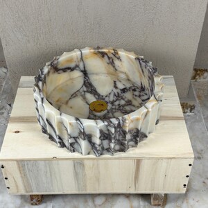 May include: A marble vessel sink with a unique, textured rim. The sink features a blend of cream, brown, and gray tones, with a gold-colored drain. It is set on a light-colored wooden base, showcasing the natural veining of the stone.
