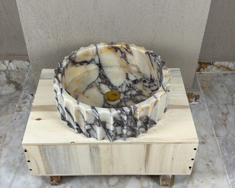 May include: A marble vessel sink with a unique, textured rim. The sink features a blend of cream, brown, and gray tones, with a gold-colored drain. It is set on a light-colored wooden base, showcasing the natural veining of the stone.