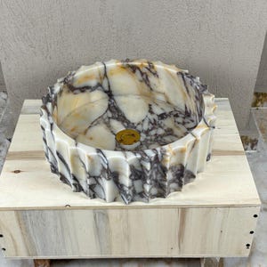 May include: A decorative marble sink with a unique, ridged exterior. The sink features a circular basin with a gold-colored drain cover. The marble has a cream and gray veined pattern. The sink is displayed on a light-colored wooden box.