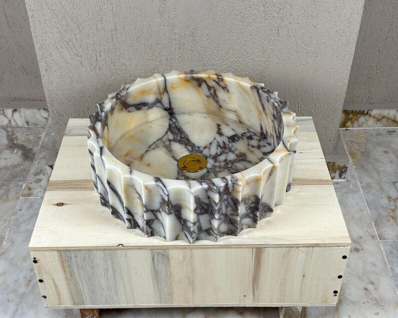 May include: A decorative marble sink with a unique, ridged exterior. The sink features a circular basin with a gold-colored drain cover. The marble has a cream and gray veined pattern. The sink is displayed on a light-colored wooden box.