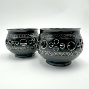 Może przedstawiać: Dwie ciemnoszare ceramiczne misy z błyszczącym wykończeniem. Każda misa ma szeroki, zaokrąglony korpus z podstawą oraz ozdobny pasek z wytłoczonymi kółkami u góry i u dołu, z większymi wyciętymi otworami w kształcie kół pośrodku.