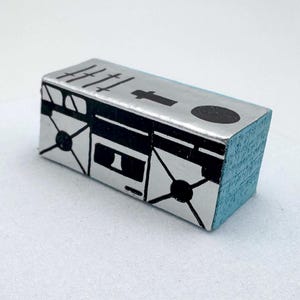 May include: A rectangular wooden block stamp with a light blue painted side and a silver top surface. The silver surface features a black graphic print of a boombox, including speakers, a cassette deck, and tally marks along the top edge.