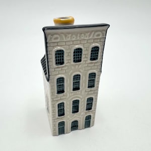 Może przedstawiać: Ceramiczna miniaturowa figurka w kształcie wysokiego, wąskiego domu o fakturze jasnoszarej cegły, z ciemnoszarymi łukowatymi oknami i czarnym wykończeniem dachu. Z góry konstrukcji wystaje mały, zaokrąglony żółty komin.