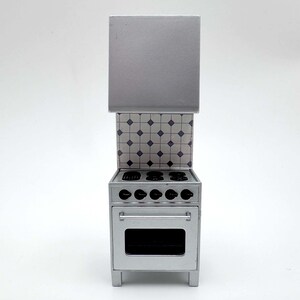 May include: A miniature silver kitchen stove featuring a tall hood, a backsplash with a white and blue diamond tile pattern, five black circular burners on the cooktop, five black control knobs, and an oven door with a silver handle.