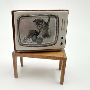 May include: A miniature television set with a brown frame and white front panel featuring a black and white graphic of a kitten playing with yarn on the screen, resting on a small, light brown wooden table with four thin legs.