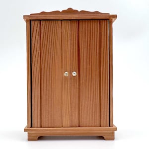 May include: A small, rectangular wooden cabinet with a natural light brown wood grain finish. The cabinet features two vertical doors, each equipped with a single small, round, metallic knob. The top has a decorative arched trim.
