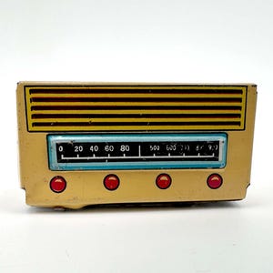 Könnte beinhalten: Ein rechteckiges Blechspielzeug-Radio in Cremefarben mit einem schwarz-gelb gestreiften Lautsprechergitter. Die Vorderseite zeigt eine hellblaue Radioskala mit weißen Zahlen von 0 bis 900 und vier kreisförmige rote Bedienknöpfe.