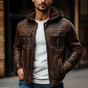 Op de afbeelding: Bruine leren jas met capuchon en twee voorzakken. De jas heeft een ritssluiting en een distressed afwerking. Het model draagt een wit t-shirt en een blauwe jeans. Een jas in klassieke stijl.