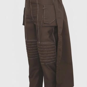 May include: Brown pants with white stitching and a unique design. The pants feature multiple pockets and quilted knee panels. A long, flowing panel extends from the waist. The pants are displayed on a mannequin.