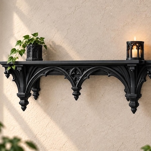 Può includere: Una mensola da parete nera caratterizzata da intagli ad arco acuto e terminali decorativi. La mensola sostiene una piccola pianta verde in vaso sul lato sinistro e una candela bianca accesa in un portacandele nero abbinato sulla destra.