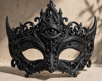 Könnte beinhalten: Eine schwarze, verzierte Maske für einen Maskenball mit einem augenförmigen Design. Die Maske weist komplizierte Schnörkel, Hörner und ein zentrales Augenmotiv auf. Die Maske besteht aus einem dunklen Material und ist so konzipiert, dass sie die Augen und die Nase bedeckt.