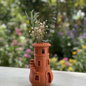 Puede incluir: Un jarrón decorativo de color terracota con forma de faro, con ventanas arqueadas y una pequeña escalera. El jarrón contiene flores secas y vegetación. El fondo es una vista borrosa de un jardín.