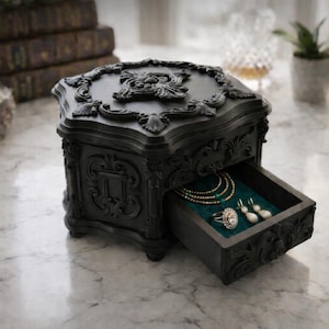 Puede incluir: Una caja de joyería negra y ornamentada con un cajón abierto que revela collares, un anillo y aretes sobre un forro de terciopelo turquesa. La caja presenta tallas detalladas y una parte superior decorativa.