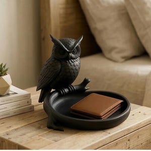 Puede incluir: Una bandeja decorativa negra con forma de búho con una cartera de cuero marrón en su interior. El búho está posado en una rama sobre la bandeja. Una pequeña planta suculenta y libros están sobre una superficie de madera.