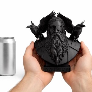 May include: A black bust of a bearded figure with an eye patch, flanked by two ravens. The bust is held in two hands, with a silver can to the left. The bust has intricate details and is on a small base.