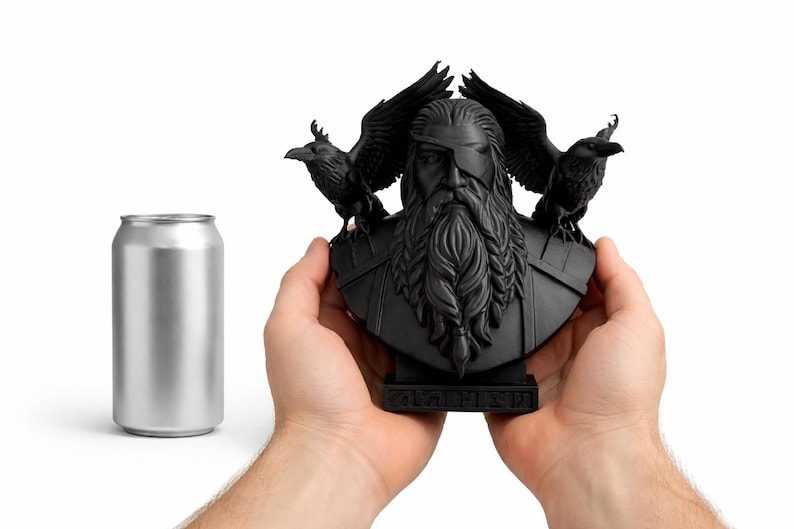 May include: A black bust of a bearded figure with an eye patch, flanked by two ravens. The bust is held in two hands, with a silver can to the left. The bust has intricate details and is on a small base.