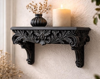 Puede incluir: Estante de pared negro y ornamentado con intrincadas tallas florales. Un pequeño jarrón negro con flores secas y una vela crema encendida se encuentran en el estante. El estante está sostenido por soportes decorativos.