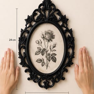 K&ouml;nnte beinhalten: Ein ovaler schwarzer Bilderrahmen mit kunstvollen Schn&ouml;rkeln und floralen Details, der eine schwarz-wei&szlig;e botanische Illustration einer Rose enth&auml;lt. Der Rahmen misst 24 cm in der H&ouml;he und 17 cm in der Breite, wie durch die Messlinien angezeigt.