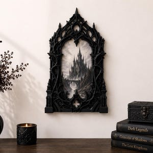 Puede incluir: Un marco de pared negro, ornamentado y arqueado, con detalles intrincados y puntiagudos de estilo gótico. El marco encierra una ilustración monocromática de un castillo imponente sobre un acantilado rocoso, un puente y un río.