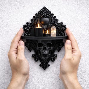 Puede incluir: Estante de pared decorativo negro con dise&ntilde;o de calavera. El estante contiene dos velas encendidas y un cristal. El estante est&aacute; sostenido por dos manos sobre un fondo blanco texturizado. El estante tiene detalles ornamentados.