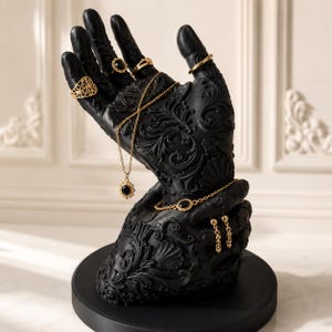 May include: A black hand-shaped jewelry display stand with intricate floral patterns. Gold-colored rings, a necklace, bracelet, and earrings with black gemstones adorn the fingers and wrist. The stand sits on a round black base.
