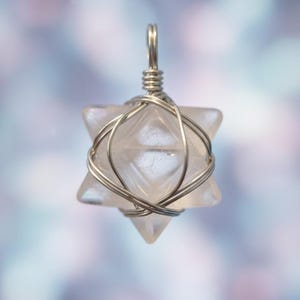 May include: A clear, star-shaped pendant with a silver wire wrap. The pendant has a six-pointed star design, with a loop at the top for hanging. The background is a soft, blurred mix of colors.