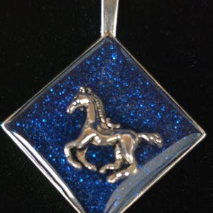 May include: Silver-toned pendant with a running horse design. The pendant has a diamond shape with a sparkly blue background. The horse is in the center of the pendant. The pendant has a silver bail.