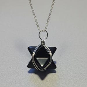 May include: A black star-shaped pendant necklace with a silver chain. The pendant is wire-wrapped and features a geometric design. The necklace is displayed against a light gray background.