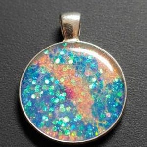 May include: Round pendant with a silver-colored bezel. The pendant features a colorful design with blue, orange, and green glitter set in a clear resin. The pendant is displayed against a dark background.