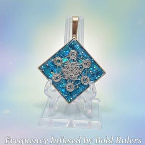 May include: A pendant with a gold-colored bail and a square, teal-colored body. The body is filled with glitter and features a silver-colored, geometric design. The pendant is displayed on a clear stand. The text "Frequency Infused by Bold Rulers" is at the bottom.