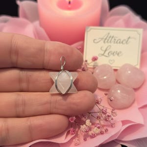 May include: A star-shaped rose quartz pendant wrapped in silver wire. The pendant is held in a hand, surrounded by pink rose petals, rose quartz stones, a lit pink candle, and a card that says "Attract Love".