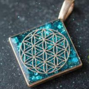 May include: Square pendant with a gold-colored frame and bail. The pendant features a teal-colored background with glitter and a gold-colored Flower of Life design in the center. The pendant is displayed on a dark, textured surface.