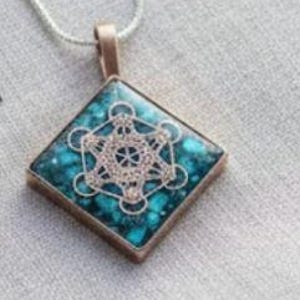 May include: A square pendant with a turquoise-colored inlay and a metallic geometric design. The pendant is set in a bronze-colored metal frame and hangs from a silver chain. The design features circles and lines.