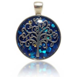May include: A silver-toned pendant with a circular, blue-glittered background. A silver tree design is centered on the pendant. The pendant has a silver loop at the top for a chain. The pendant is likely for a necklace.