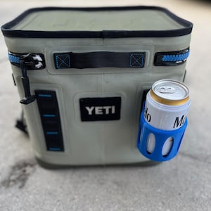 Op de afbeelding: Een zachte, lichtgroene koeler met een zwarte rits en handvat, met het merk "YETI". Een blauwe blikhouder houdt een bierblikje vast. De koeler is ontworpen om drankjes koud te houden.