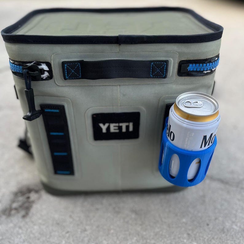 Op de afbeelding: Een zachte, lichtgroene koeler met een zwarte rits en handvat, met het merk "YETI". Een blauwe blikhouder houdt een bierblikje vast. De koeler is ontworpen om drankjes koud te houden.