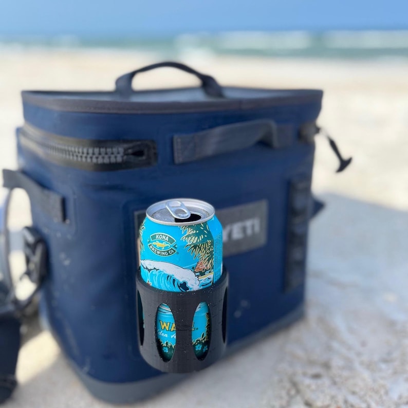 Op de afbeelding: Een marineblauwe YET cooler met een zwarte rits en handvat staat op het zand. Een zwarte bekerhouder houdt een Kona Brewing Co. drankblikje met een tropisch ontwerp vast.