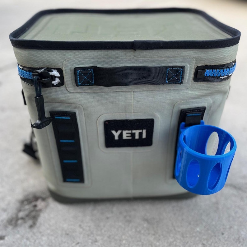 Op de afbeelding: Een zachte, lichtgrijze koeler met zwarte rand en blauwe rits. Het heeft een zwarte handgreep en een blauwe bekerhouder. De merknaam "YETI" is zichtbaar op de voorkant.