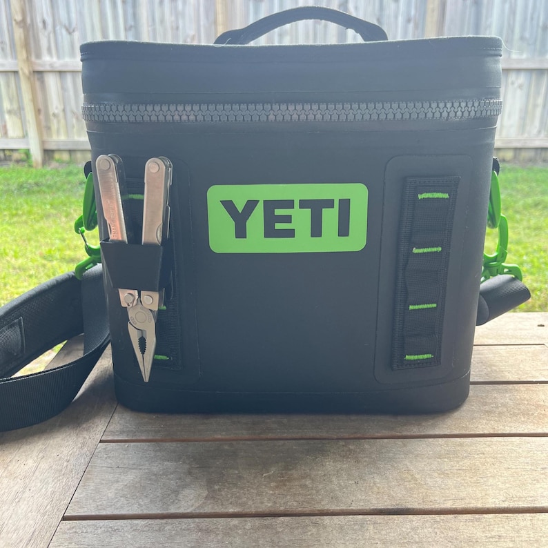 May include: Black insulated cooler bag with a green "YETI" logo. The bag has a black shoulder strap, a handle, and a side pocket holding a multi-tool. Green accents are visible on the bag's exterior.