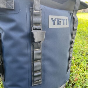 May include: Close-up of a navy blue cooler bag with the brand name "YETI" in gray. The bag has black straps and a black buckle closure. The background is blurred, showing green grass.