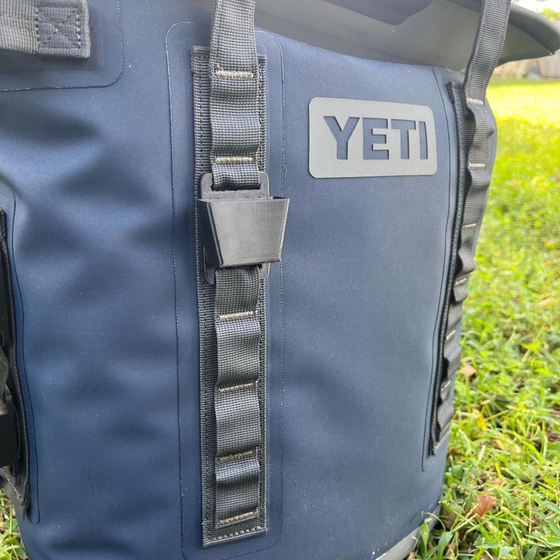 May include: Close-up of a navy blue cooler bag with the brand name "YETI" in gray. The bag has black straps and a black buckle closure. The background is blurred, showing green grass.